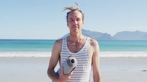 Retrato-En-Video-De-Un-Hombre-Caucásico-Sonriente-Con-Rastas-Sosteniendo-Una-Estera-De-Yoga-En-Una-Playa-Soleada