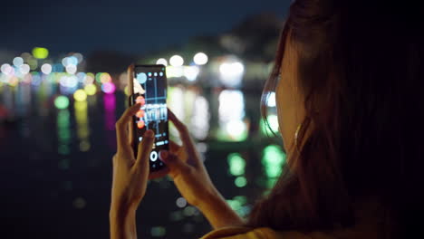 Asian-woman-takes-photos-of-night-river-reflections-in-Hoi-An,-Vietnam-with-blurred-background