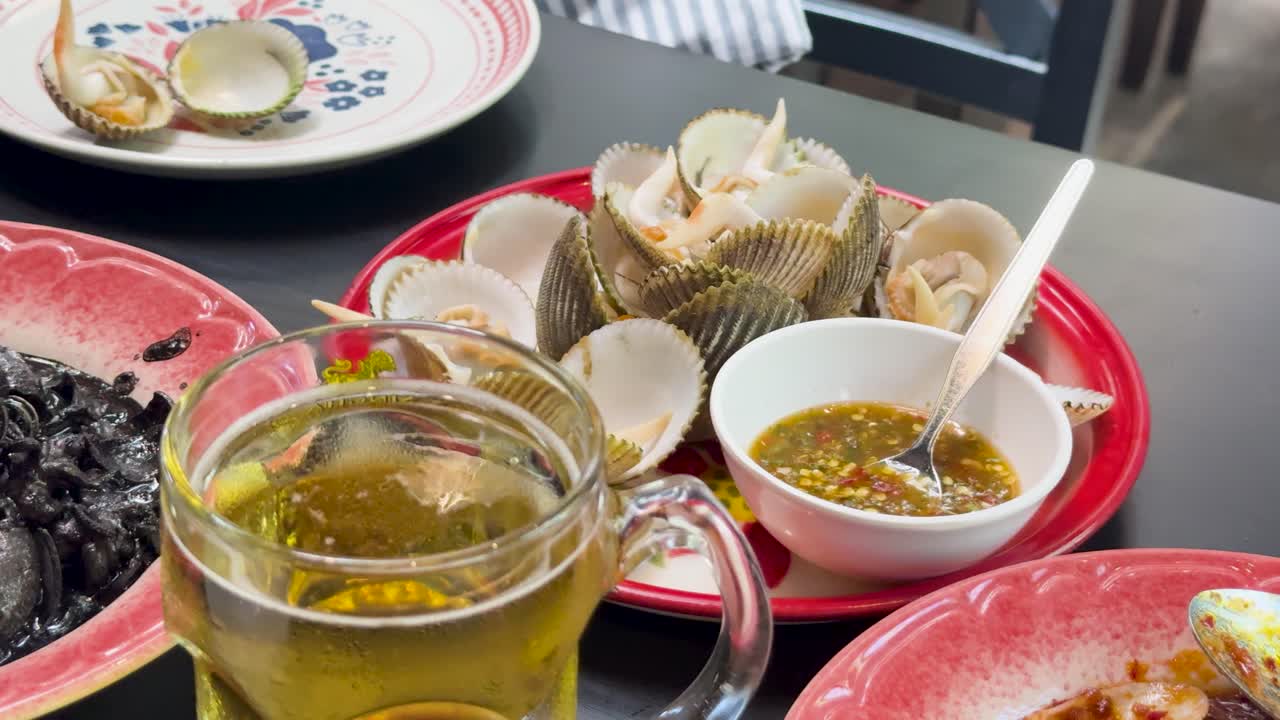 A close-up sequence shows blood cockles, spicy dipping sauce, and a glass of beer on a restaurant table under bright indoor lighting, with subtle camera movement