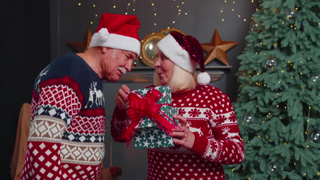 Grandfather-gifting-Christmas-present-box-to-surprised-grandmother,-happy-senior-couple-in-hats