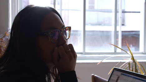 An-asian-woman-thinking-about-something-as-she-sits-at-her-laptop-computer-working-from-her-apartment