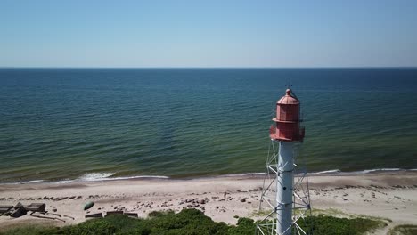 beautiful aerial view of white painted steel lighthouse with red top located in pape, latvia at baltic sea coastline in sunny summer day, distant sea, wide angle drone shot moving forward slow