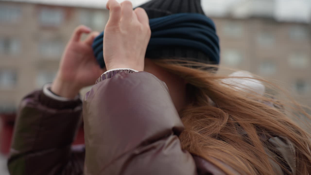 여성이 쌀쌀한 도시 환경에서 비니를 고쳐 쓰고 있어요. 도시의 냉기 속에서 여성의 모자를 다듬는 모습이 클로즈업되었고, 여성이 차가운 바람 속에서 집중된 시선으로 니트 모자를 조심스럽게 정돈하고 있어요.
