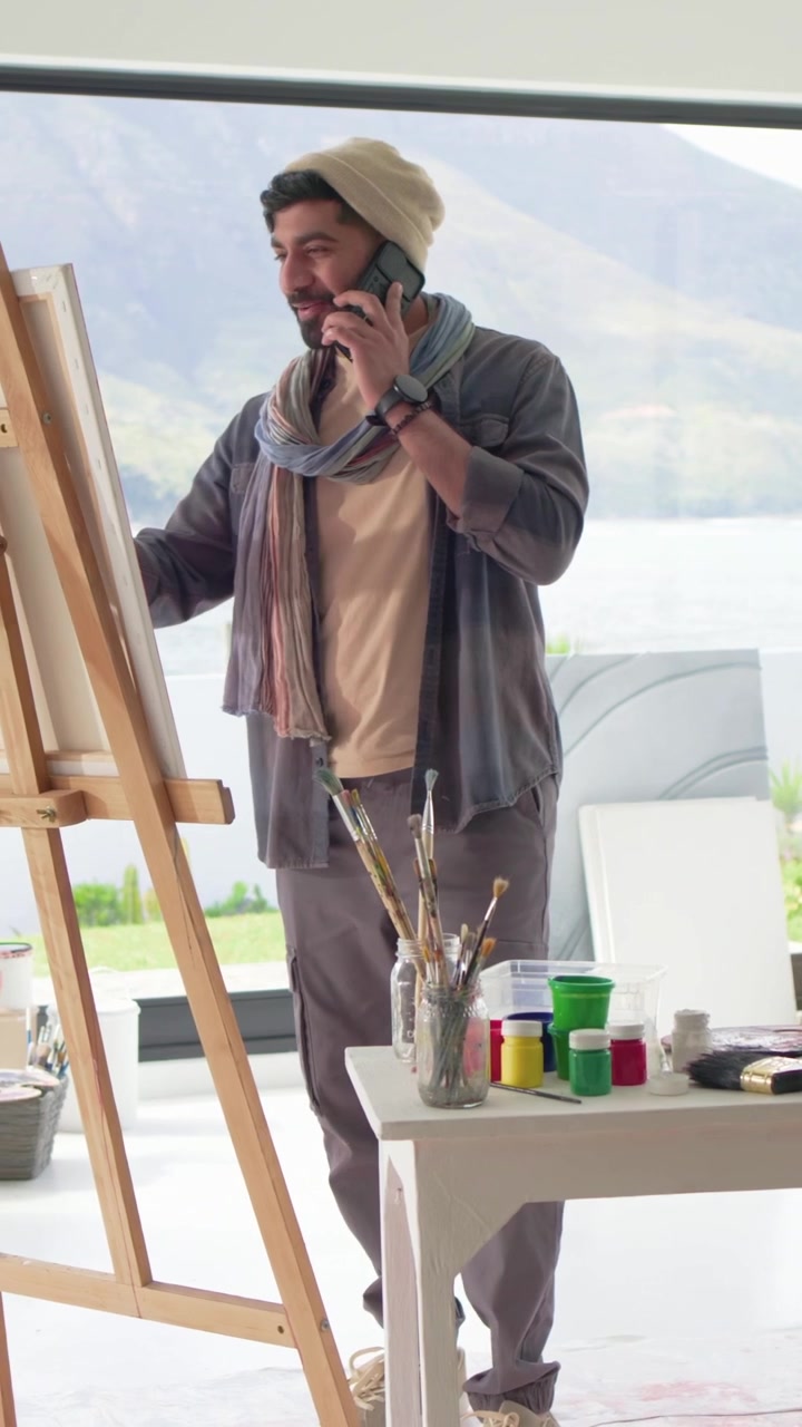 Vertical video: Painting artist in scarf taking phone call, using brushes at easel by lake