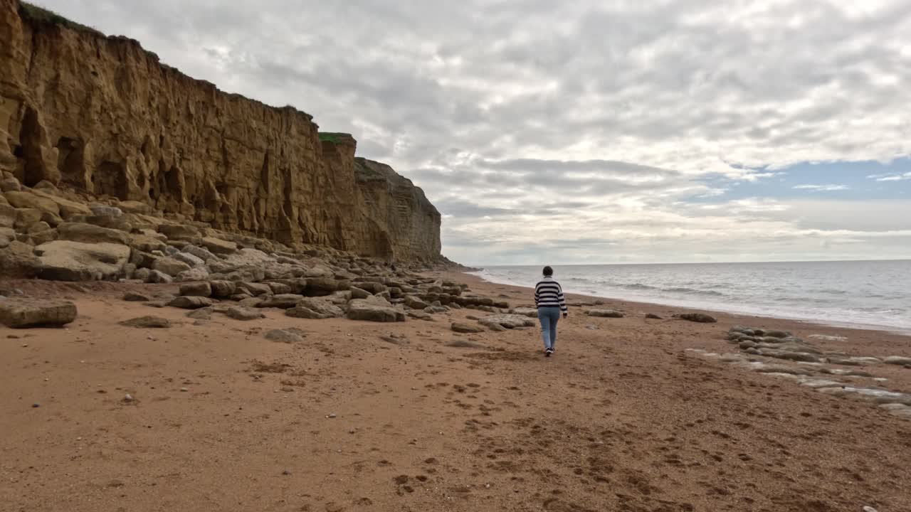 A women walking along a Jurassic Coast beach in Dorset. The camera moves sideways to the right as the female walks away along the beach wide the sea on the right and the cliffs left.