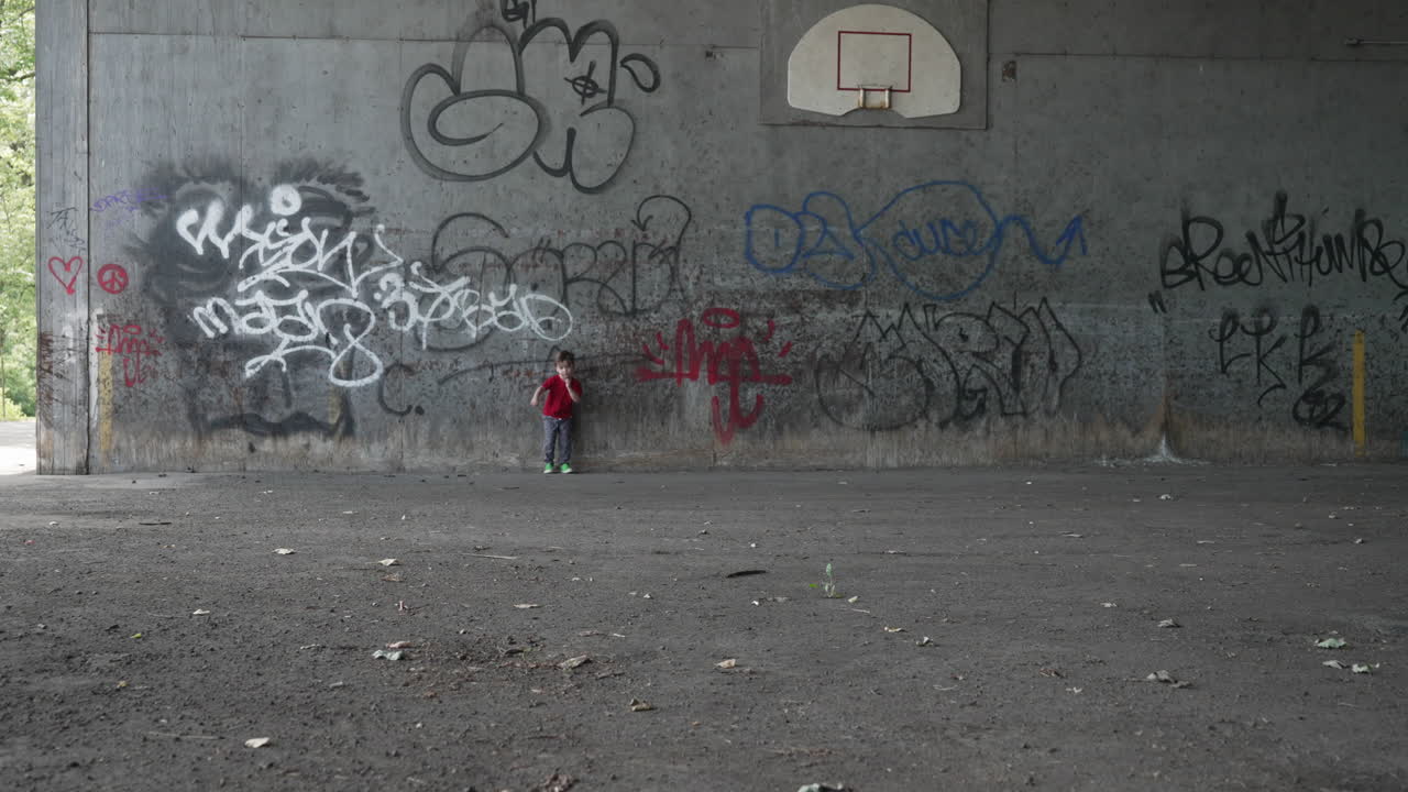 Video al rallentatore di un bambino piccolo con una maglietta rossa appoggiato a un muro graffitato che corre verso la telecamera