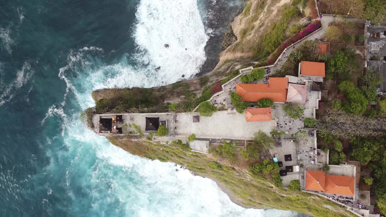 4K top-down drone shot captures the iconic Uluwatu cliffside Temple complex and in Bali. Powerful ocean waves crash against the sheer rock face