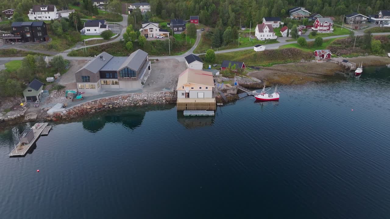 Aerial drone view of a new wooden pier and boathouse in Tromsø, Norway. Features a docked fishing boat, residential houses, and green hills along the scenic fjord coastline