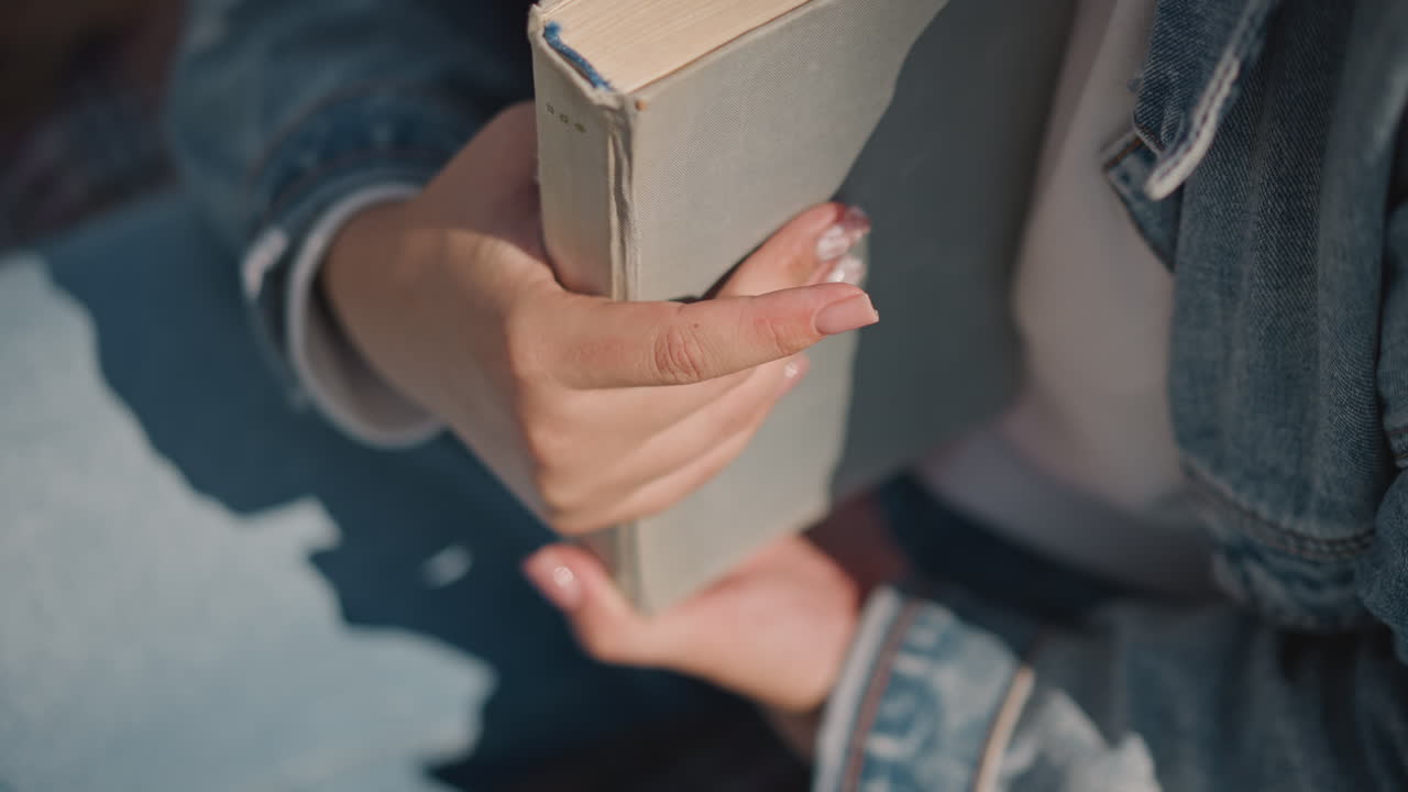 Jovem mulher segurando livro no colo, jaqueta jeans e unhas feitas banhada pela luz solar quente, close-up mostra capa dura texturizada, pegada suave, postura relaxada em banco ao ar livre, momento de estudo tranquilo de fim de semana