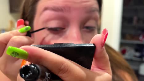 close up of young woman putting mascara on eyelash while holding small portable mirror