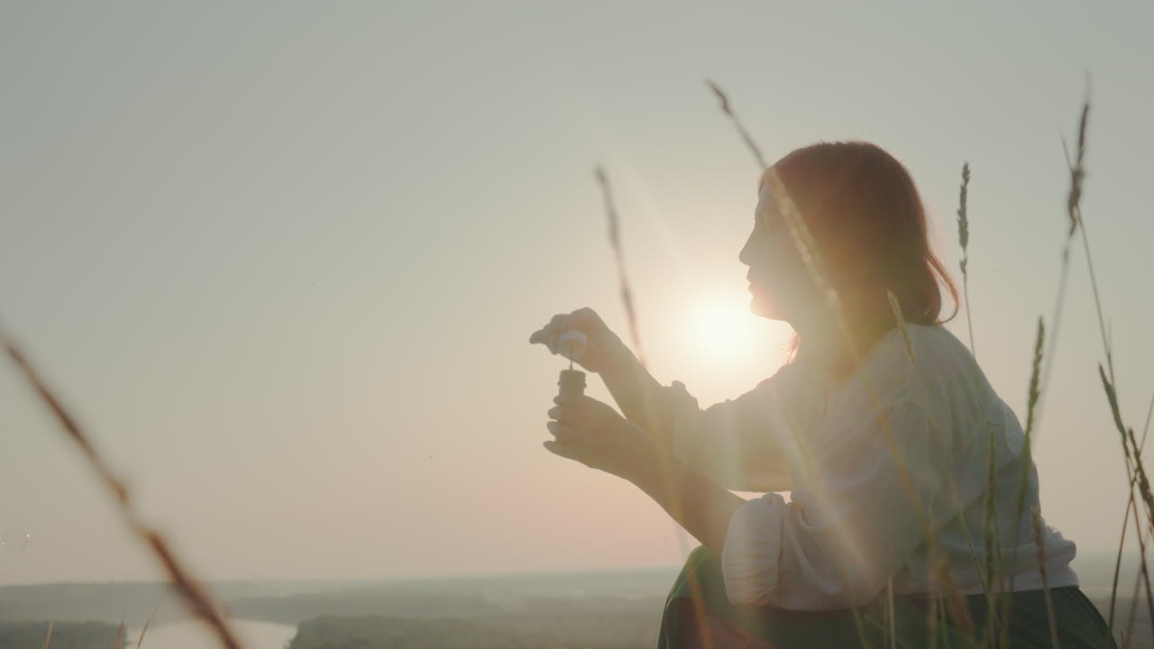 Woman in soft silhouette blowing bubbles in peaceful outdoor setting, surrounded by tall grain stalks and backlit by warm sun glow, with drifting bubbles and serene river valley fading into distance