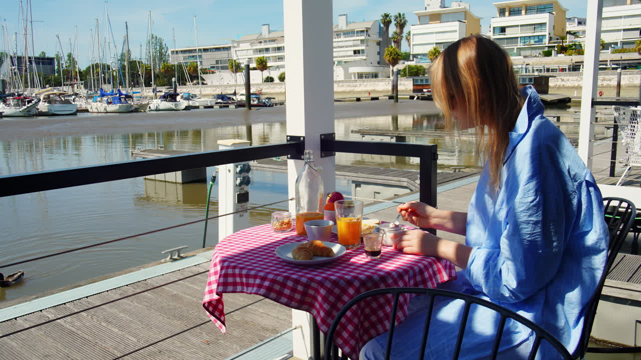 Mujer disfrutando del desayuno en una terraza exterior con vistas a una marina.