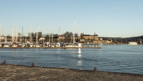 People-walking-Oslo-harbour-at-sunset,-with-Akershus-Fortress-behind,-time-lapse