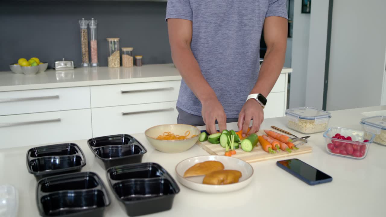 Afroamerikanischer Mann in blauem T-Shirt, der auf einer weißen Kücheninsel schneidet und Meal-Prep-Behälter befüllt.