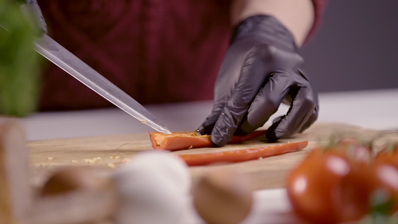 Slow-motion of a chef preparing a red chili pepper for slicing. The sharp knife is ready to cut, with black gloves ensuring hygiene. Fresh ingredients like tomatoes and herbs add depth to the scene