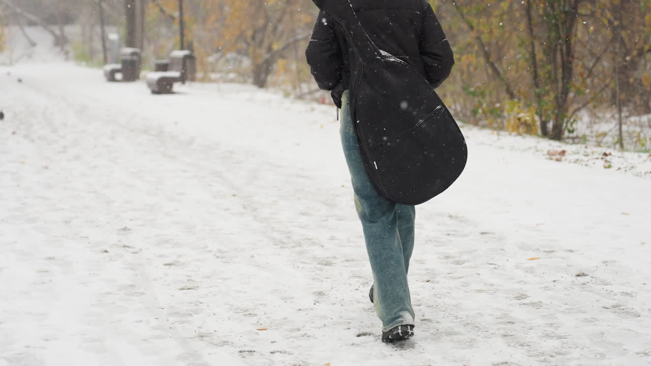 vista trasera de alguien con gorra negra y sudadera con capucha con las manos en el bolsillo, llevando una guitarra en el paquete, caminando en la nieve, banco y árboles en el fondo con una paloma en la distancia