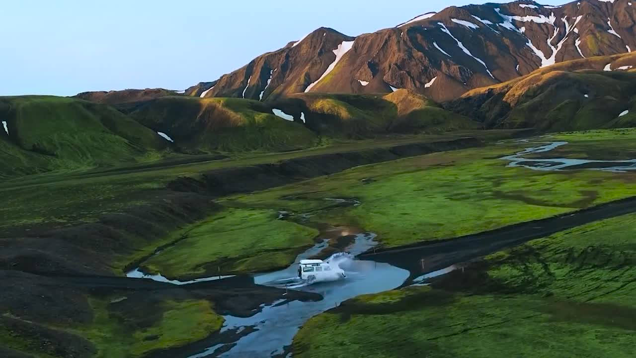 Aerial drone flying besides a white 4x4 truck vehicle driving through cold fresh river water in green mossy and grassy landscape in the Greenland or Iceland mountains during sunny day with snow seen.