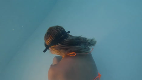 cinematic shot of a woman in an orange bikini and vintage diving goggles moving gracefully underwater in clear blue waters shot in 4k, 120 fps, slomo