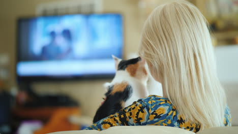 blonde woman with a kitten in her arms is watching tv rear view