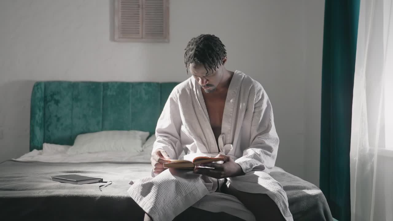Man sitting on bed reading book, morning light coming through window. Calm atmosphere, wearing white bathrobe, focused on reading. Cozy room, peaceful moment captured