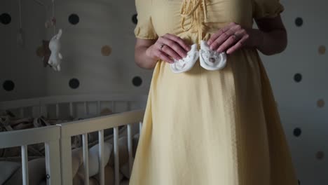 pregnant woman in yellow dress standing next to wooden crib in nursery showing cut little white socks for the baby