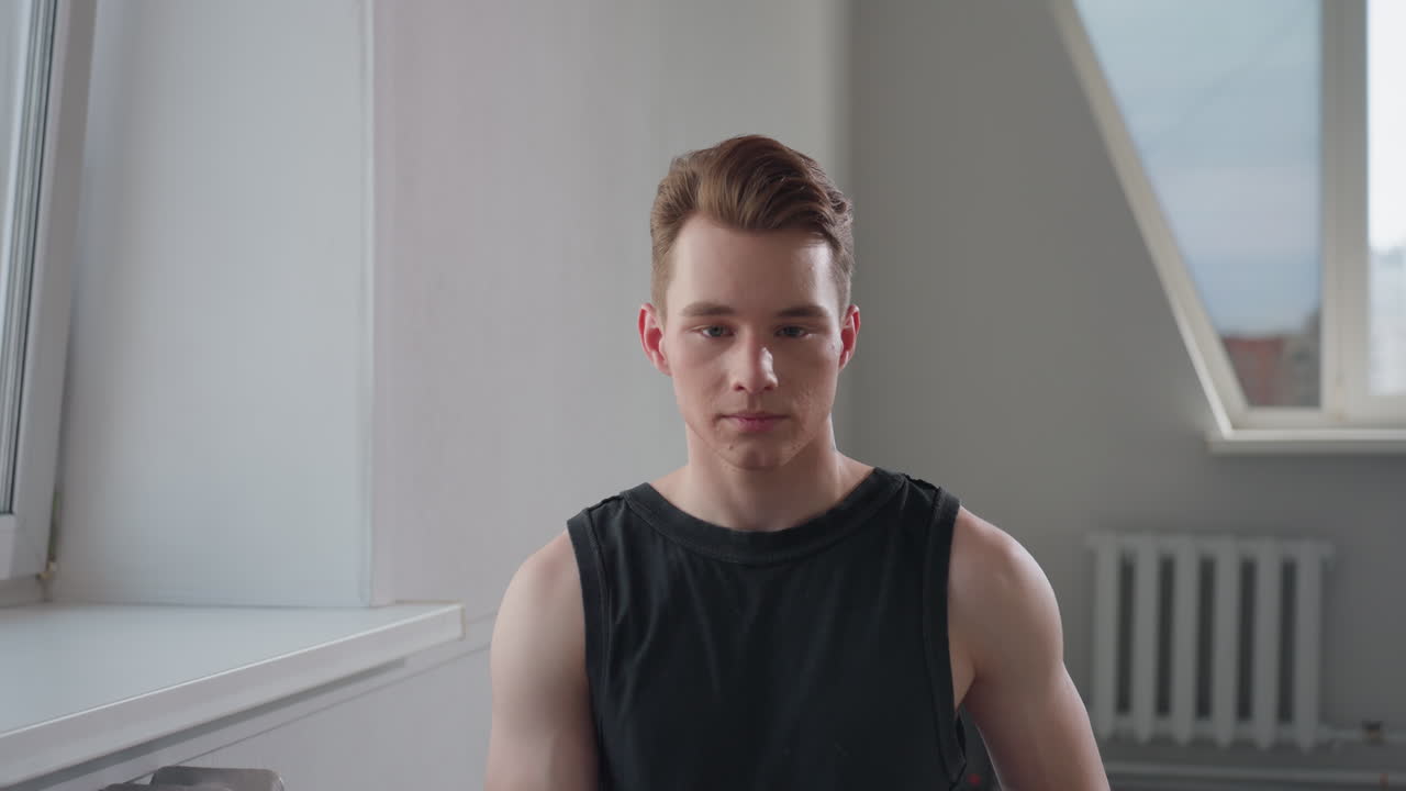 Young man in black sleeveless top sits on stool in empty bright room by large window, gazes forward with calm focused expression, hands resting on thighs