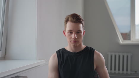 young man in black sleeveless top sits on stool in empty bright room by large window, gazes forward with calm focused expression, hands resting on thighs