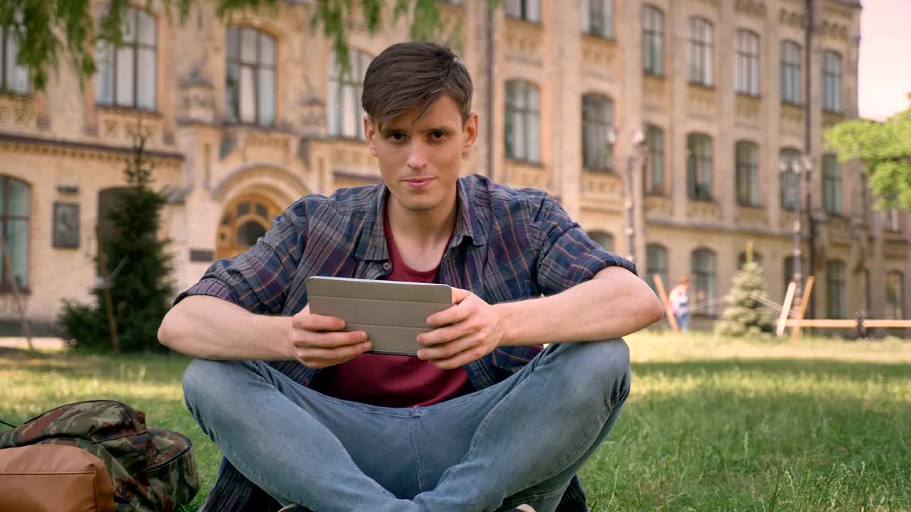 젊은 잘생긴 남자가 공원의 잔디에 앉아, 태블릿에서 인터넷 페이지를보고, 카메라를보고, 휴식 개념, 의사소통 개념, 배경에 기반을 두고 있습니다.