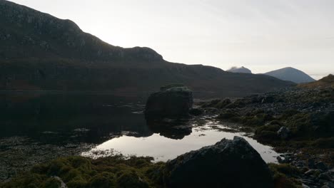 Suaves-Ondas-Se-Mueven-A-Través-De-La-Superficie-Del-Agua-Oscura-Y-Tranquila,-Charcos-De-Marea-Y-Algas-Marinas-En-Un-Lago-Marino-En-Escocia-Mientras-El-Sol-Se-Pone-Detrás-De-Las-Montañas