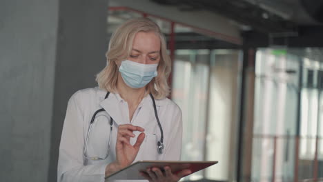 a blonde, middle aged female doctor in a facemask consults data on a tablet 1