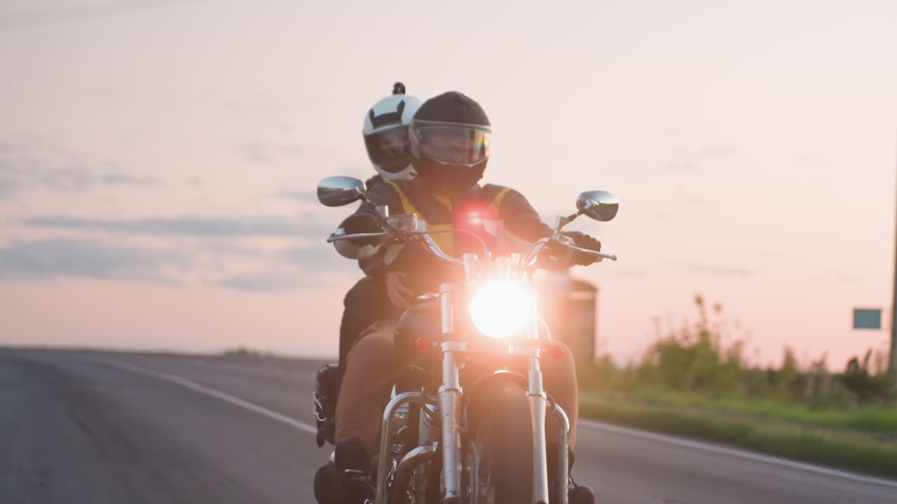 Racer wearing helmet rides motorbike with passenger during evening, headlight shining while car travels past on opposite lane, soft pastel sky above countryside road