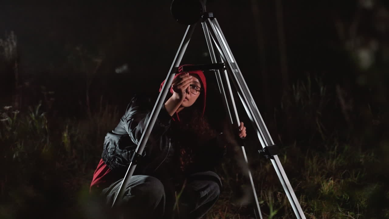 astrofilo amatoriale in un campo di notte che regola il telescopio accovacciato dietro il treppiede, indossa un berretto rosso e una giacca scura, spiazzo erboso sotto deboli stelle, regolazione attenta dell'oculare e paziente scansione del cielo