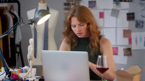 female owner of fashion business with glass of wine working late on laptop in studio