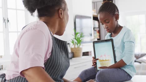 Happy-african-american-grandmother-and-granddaughter-baking-in-kitchen,-using-tablet,-copy-space