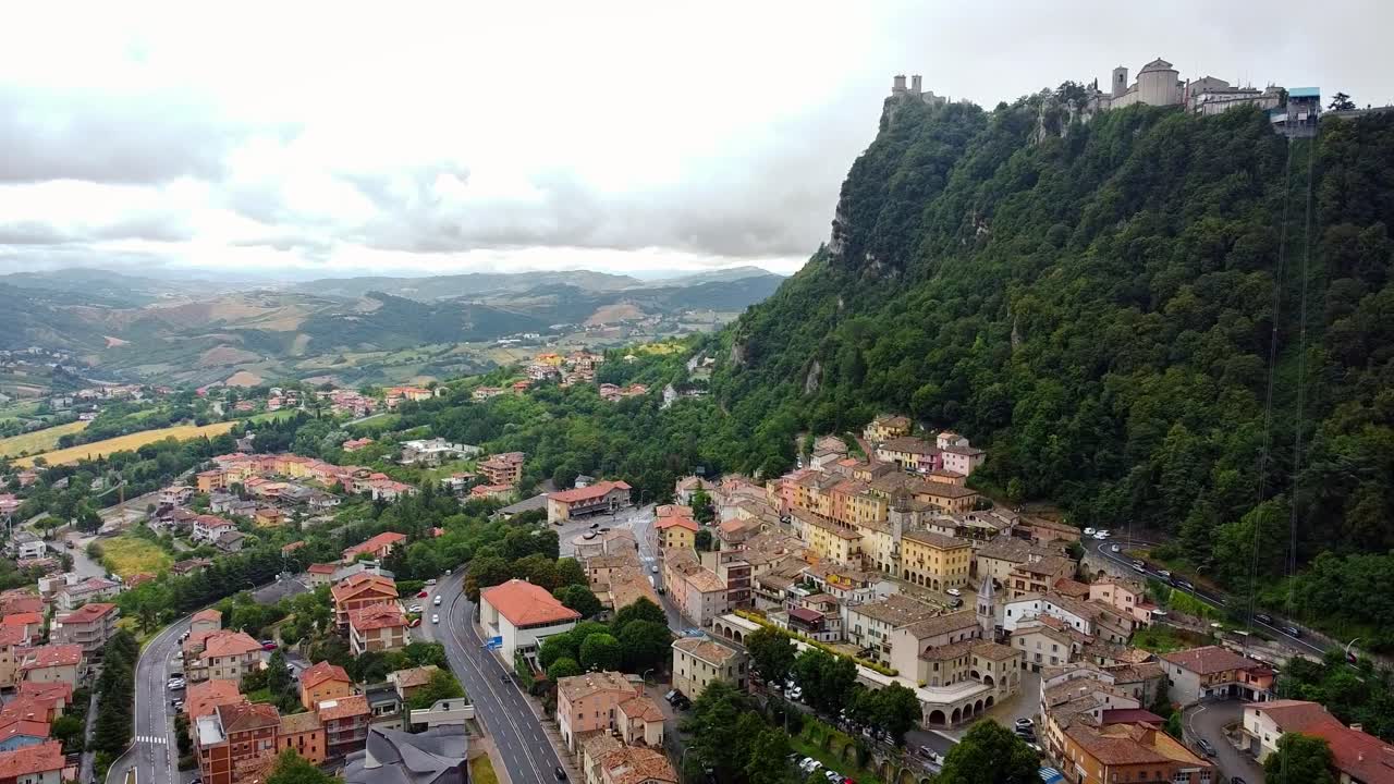 Aerial view pulling back over San Marino castle, old town buildings, forested hills and cable car, scenic mountain backdrop with historic charm