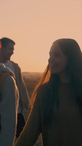 happy young couples girls and guys walk along meadow above river to modern campsite in calm autumn evening at back sunset