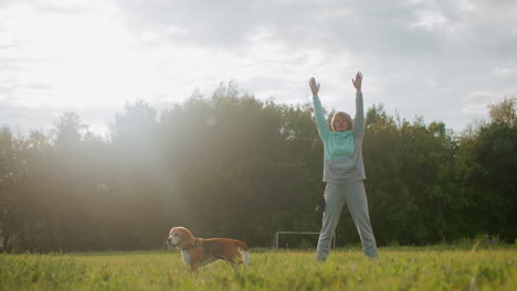female coach wearing mint tracksuit standing on open grassy park stretching arms outward under cloudy sky with beagle dog beside during peaceful morning workout session promoting fitness