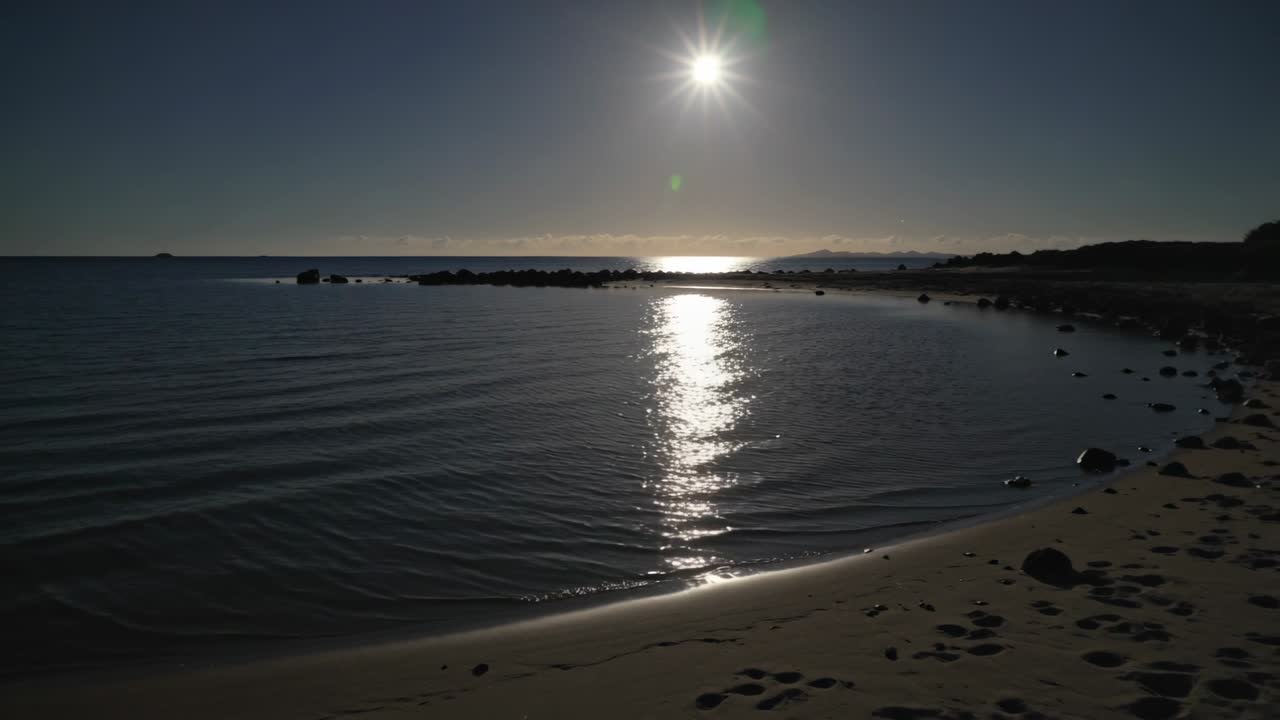 Superfície do mar ondulando, projetando reflexo solar vertical na costa, brisa empurrando ondulações para além das rochas.