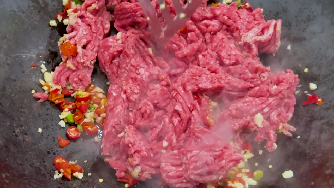 Raw minced meat is stirred and broken apart with a spatula in a hot wok, mixing with chopped spicy vegetables under bright, natural lighting