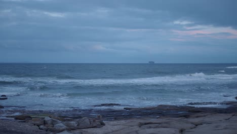 Jenseits-Des-Felsigen-Strandes-Segelt-Ein-Großes-Frachtschiff-Am-Horizont-Der-Küste,-Während-Die-Untergehende-Sonne-Alles,-Was-Sie-Berührt,-Mit-Einem-Kalten-Blau-Färbt,-Was-Eine-Kühle-Nachmittagsstimmung-Erzeugt