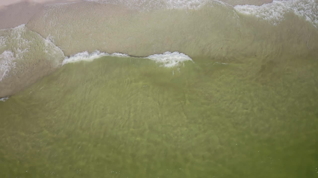 Shallow emerald water shimmers as small waves feather toward a pale shoreline, revealing rippled sandbars and drifting foam patterns that pulse beneath the surface with gentle, hypnotic motion