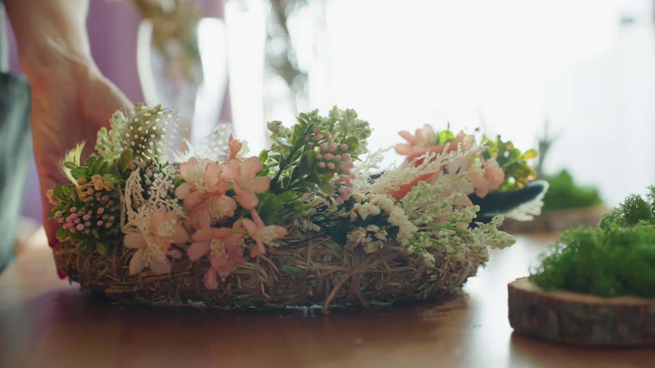 Female florist arranging handmade wreath with flowers, greenery, and rustic twigs on wooden table decorated with moss, natural light highlighting artisan craft process in cozy creative workspace