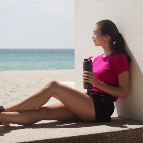 Sportliche-Frau-Mit-Flasche-Wasser-Entspannt-Nach-Dem-Training-Am-Strand-On