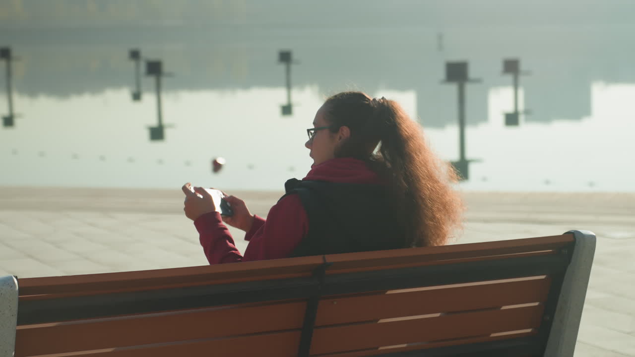 Caucasian woman on bench by waterfront scrolling phone in reflective mood, long hair haloed by backlight, distant boats and calm surface, serene golden light evokes solitude and quiet observation