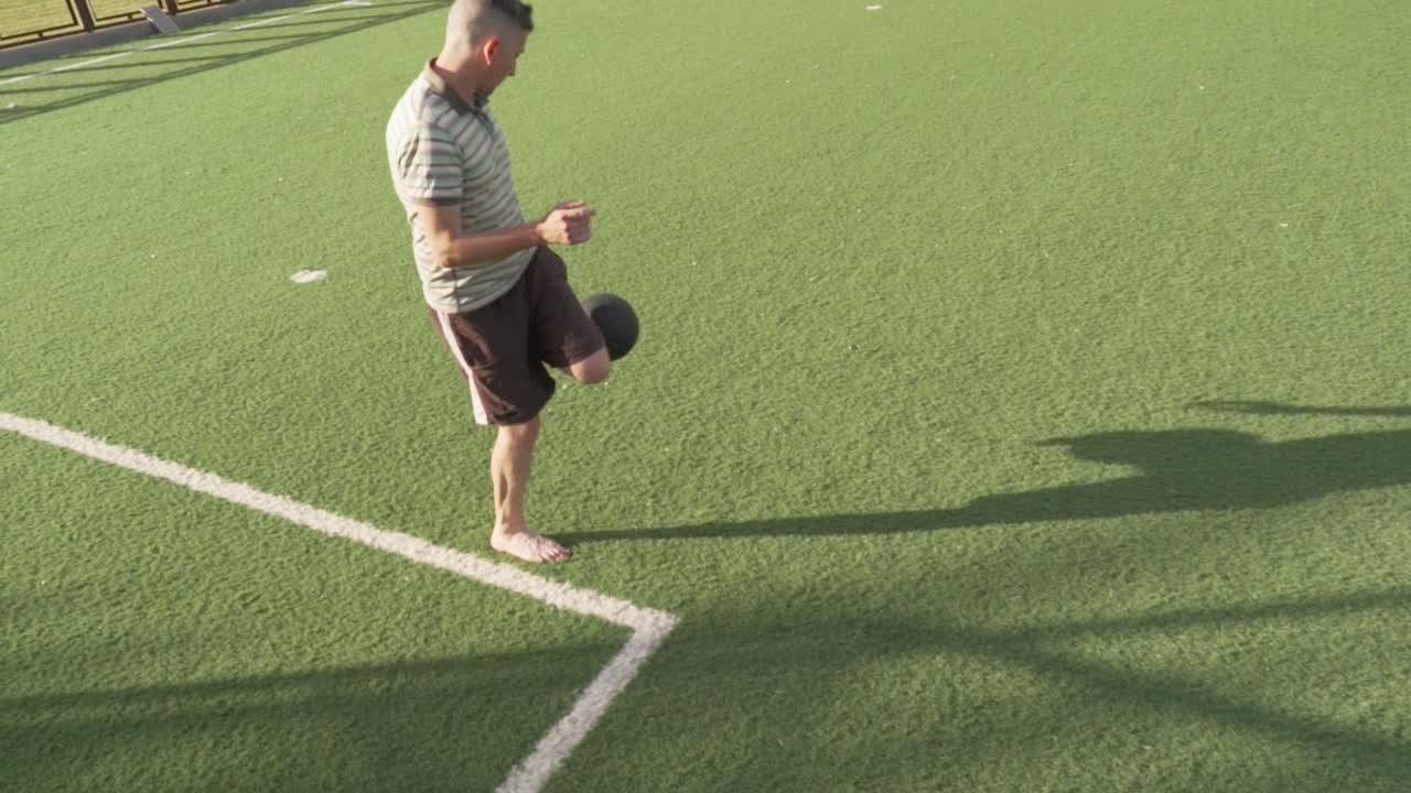 Skilled male juggling soccer ball barefoot with playful energy while standing on turf field sideline during sunny summer day
