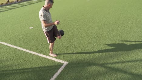 skilled male juggling soccer ball barefoot with playful energy while standing on turf field sideline during sunny summer day