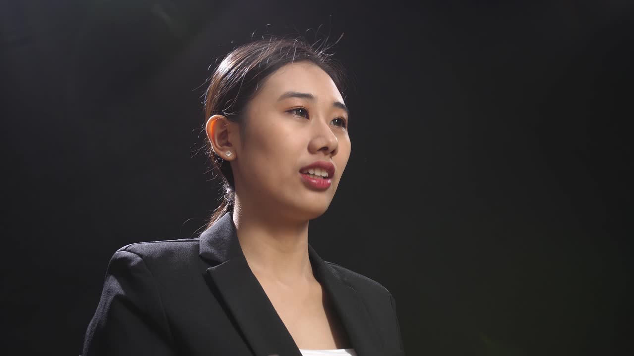 블랙 스크린 스튜디오 에서 연설 하는 동안 손 을 고 있는 비즈니스 슈트 를 입은 아시아어 연설자 여자 의 측면 시각
