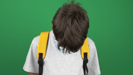 niño adolescente con máscara de protección médica y mochila bajó la cabeza y miró hacia abajo. niño de pie cerca de la pantalla verde fondo clave croma. video 4k
