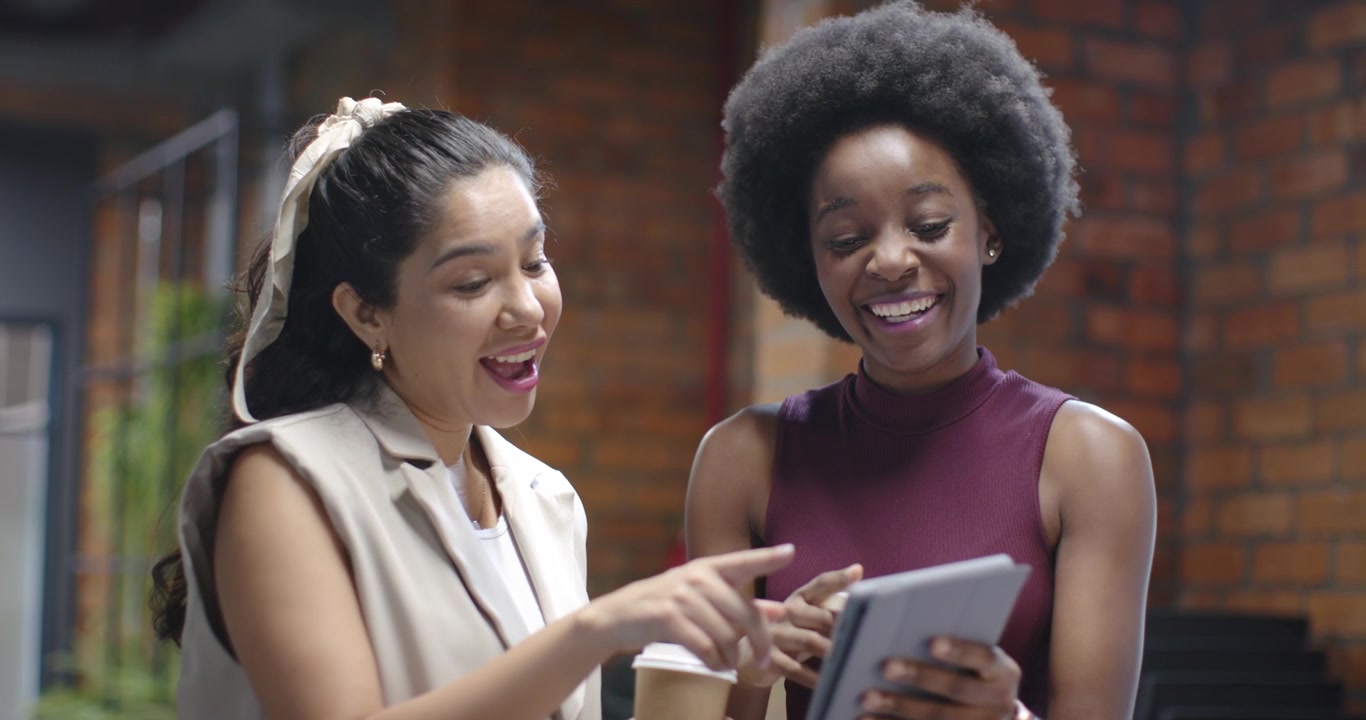 Latinamerikanske og afrikanske kvinder i loungeområde i højre side, der tapper på tablet, deler indhold og griner med kaffe