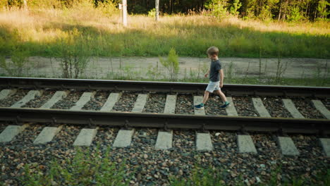 little boy walking confidently on rail track with forest and electric pole in background, surrounded by green plants and sunlight casting shadows across rocky ground in rural setting during daytime
